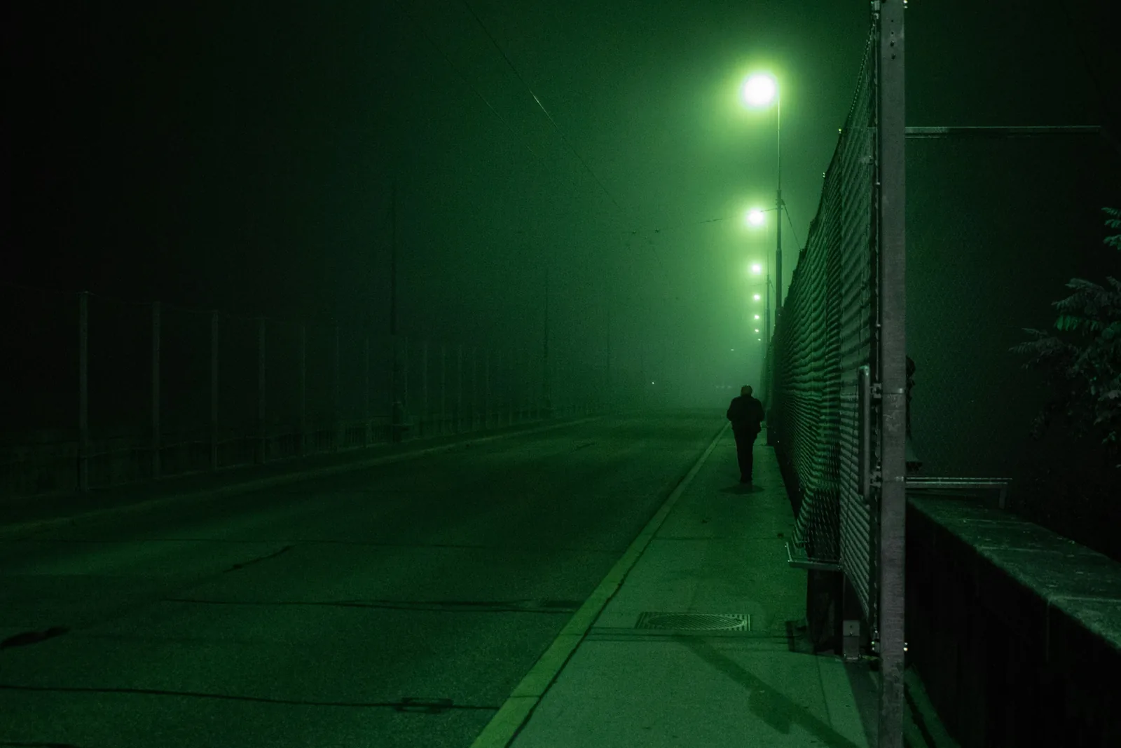 A lone figure walking away along a foggy bridge at night under green street lamps, a chain-link fence beside the pavement