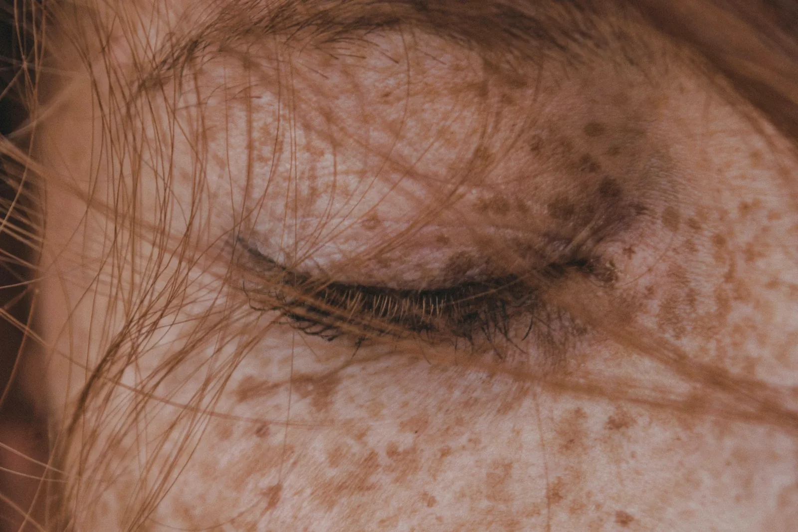 A closed eye in warm light, lashes and loose strands of hair falling across freckled skin